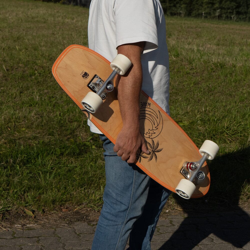 Person trägt das Surfskate Wood Wave unter dem Arm, Holzoptik mit Palmenmotiv
